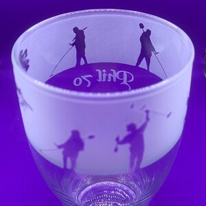 May include: A clear glass with a frosted upper section. The frosted area features silhouettes of golfers in action, with the word "Golf" in a stylized font. The glass has a flared shape and a detailed base.