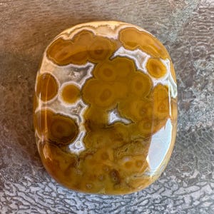 May include: A polished, oval-shaped gemstone with a unique pattern. The stone features a mix of warm colors, including shades of brown, yellow, and white. The design resembles a series of interconnected circles and cloud-like formations. The surface is smooth and reflective.