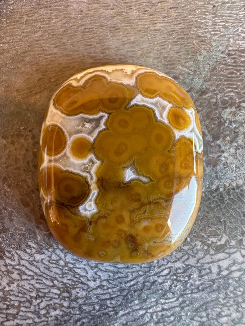 May include: A polished, oval-shaped gemstone with a unique pattern. The stone features a mix of warm colors, including shades of brown, yellow, and white. The design resembles a series of interconnected circles and cloud-like formations. The surface is smooth and reflective.