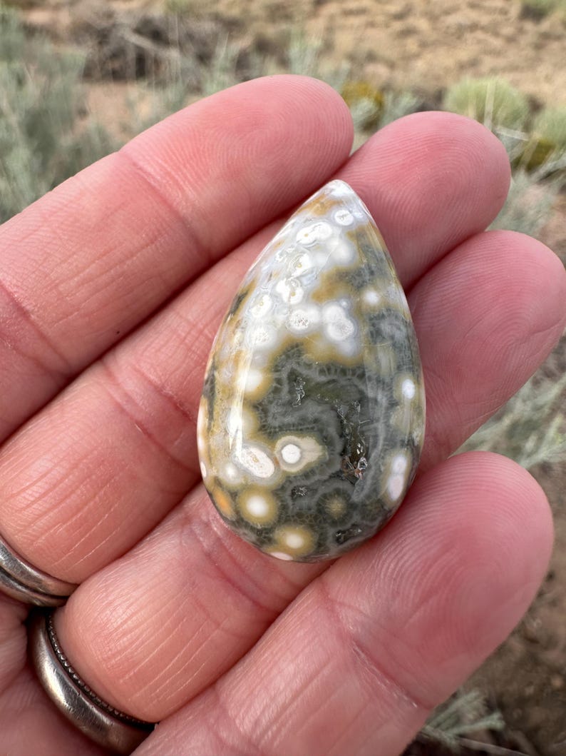 Pode incluir: Uma pedra preciosa de jaspe oce&acirc;nico polida em forma de l&aacute;grima, apresentando um padr&atilde;o complexo de orbes circulares em tons de verde oliva, amarelo mostarda e branco creme, com uma superf&iacute;cie lisa e brilhante que reflete a luz.