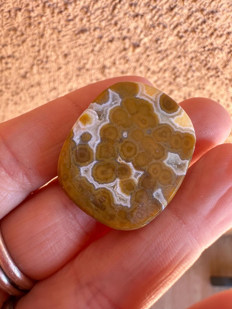May include: A polished, oval-shaped gemstone with a unique pattern of brown and tan circular designs, separated by white and cream-colored lines. The stone is held in a hand, showcasing its natural beauty and intricate details. The gemstone is a natural stone.