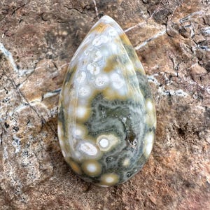 Pode incluir: Uma pedra de jaspe oce&acirc;nico polida em forma de gota, apresentando um padr&atilde;o complexo de orbes circulares em tons de verde musgo, amarelo mostarda e branco creme, com uma pequena cavidade cristalina natural vis&iacute;vel na superf&iacute;cie.