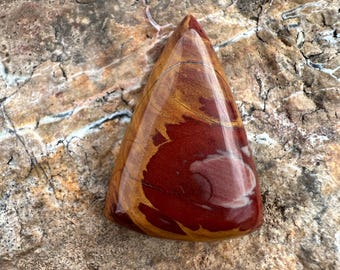 Pode incluir: Uma pedra de jaspe polida de formato triangular com uma superfície lisa e brilhante, apresentando padrões em espiral nas cores vermelho profundo, amarelo mostarda e creme. A pedra tem um topo pontiagudo e uma base arredondada.