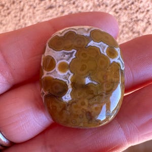 May include: A polished, oval-shaped gemstone with a unique pattern. The stone features a mix of brown, tan, and white colors, with a swirling, cloud-like design. The surface is smooth and reflective, suggesting a high polish. The gemstone is held in a hand.