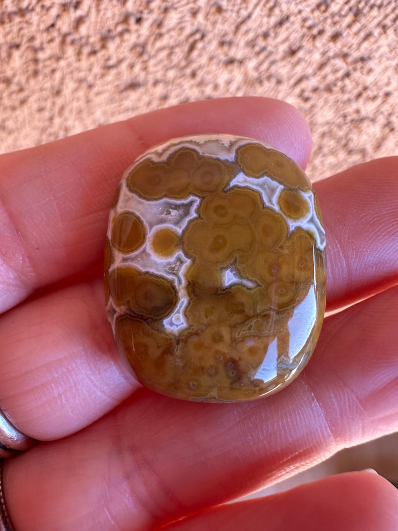 May include: A polished, oval-shaped gemstone with a unique pattern. The stone features a mix of brown, tan, and white colors, with a swirling, cloud-like design. The surface is smooth and reflective, suggesting a high polish. The gemstone is held in a hand.