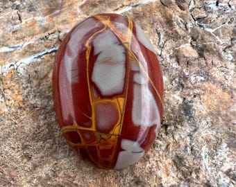 Pode incluir: Uma pedra polida de formato oval com um padrão complexo de veios em vermelho profundo, cinza suave e amarelo vibrante. A superfície é lisa e brilhante, exibindo uma rede de linhas que se cruzam, criando um aspeto de mosaico na rocha.