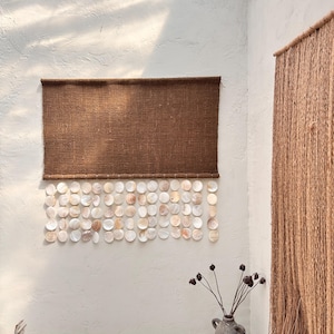 May include: A rectangular brown woven wall hanging featuring a horizontal panel of dark brown fabric with a series of circular, iridescent white shell discs suspended in vertical rows from the bottom edge, creating a layered, textured appearance.