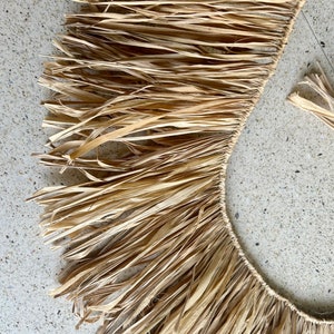 May include: A close-up of a natural fiber fringe trim, made of woven straw, with a light brown color. The fringe is arranged in a semi-circle shape.