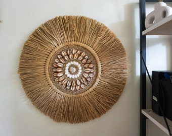 May include: A round wall hanging made of natural materials, featuring a woven base with a circular design of shells in shades of brown and white.