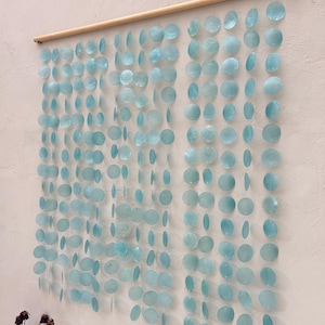 May include: A wall hanging made of light blue, round, shell-like pieces strung together on a wooden dowel rod. The hanging is made of multiple strands of shells, creating a cascading effect.