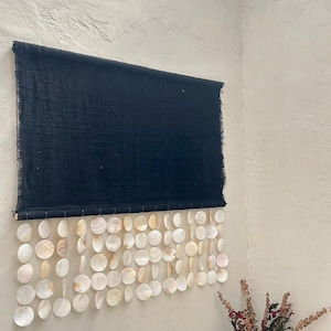 May include: A rectangular black fabric wall hanging featuring a top section of solid dark textile with frayed edges and a bottom section composed of multiple rows of circular, iridescent white capiz shells suspended from the fabric base.