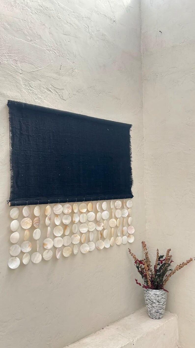 May include: A rectangular black fabric wall hanging featuring a top section of solid dark textile with frayed edges and a bottom section composed of multiple rows of circular, iridescent white capiz shells suspended from the fabric base.