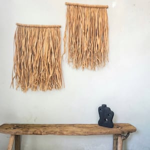 Puede incluir: Dos tapices de pared tejidos con fibras naturales cuelgan de una pared blanca. Un jarrón de cerámica negro se encuentra sobre un banco de madera rústico.