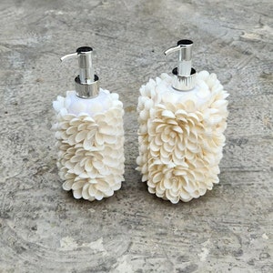 Puede incluir: Dos dispensadores de jabón de cerámica blanca con bombas plateadas. Los dispensadores están cubiertos de pequeñas conchas blancas.