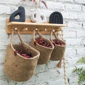 Peut inclure: Trois paniers suspendus en crochet marron avec des poignées en bois remplis de raisins rouges. Les paniers sont suspendus à une étagère en bois avec des crochets. L'étagère est fixée à un mur en briques blanches.