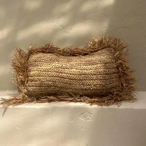 Może przedstawiać: Prostokątna poduszka wykonana z naturalnego materiału tkanego z frędzlami na krawędzi. Poduszka jest wykonana z jasnobrązowego materiału i ma fakturowaną powierzchnię.