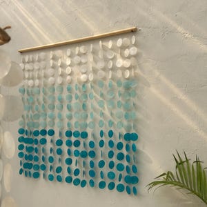 Op de afbeelding: Een wanddecoratie gemaakt van witte en blauwe, iriserende cirkels, gerangschikt in een verloop. De cirkels zijn aan een houten stok bevestigd.
