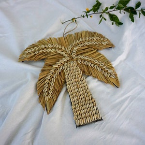 Peut inclure: Un palmier décoratif en paille naturelle et coquillages. Les frondes sont en paille tressée, et le tronc est orné de rangées de petits coquillages blancs. Une boucle en fibre naturelle est attachée pour suspendre.