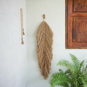 Puede incluir: Un tapiz de pared tejido de color marrón en forma de pluma. El tapiz está hecho de fibras naturales y tiene un borde con flecos. Está colgado en una pared blanca con una planta de helecho verde en primer plano.