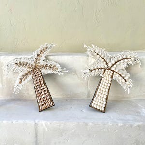 Op de afbeelding: Twee witte palmboom wanddecoraties gemaakt van schelpen en wit touw. De palmbomen bevinden zich op een witte muur.