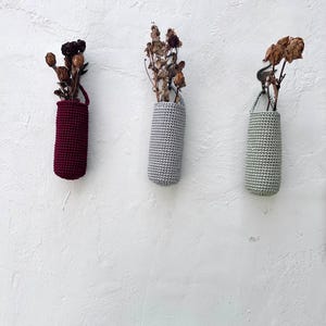 May include: Three handmade crochet vases in burgundy, gray, and sage green, each holding dried flowers. The vases are hanging on a white textured wall, showcasing a minimalist and natural aesthetic. The vases have a small loop for hanging.
