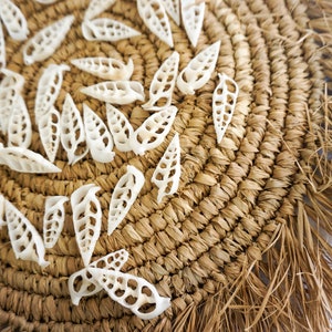 May include: A woven straw placemat with a fringe border, decorated with white seashells in the shape of leaves.