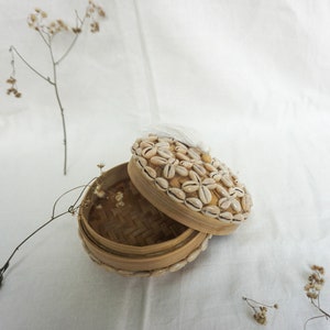 May include: A round, woven, natural-colored box with a lid decorated with white seashells. The lid has a white tassel.
