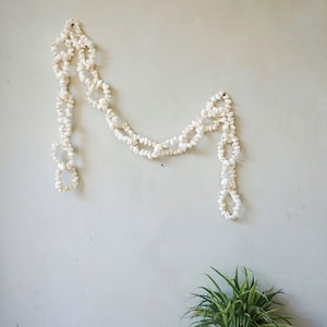 以下が含まれることがあります： 白い壁に白い貝殻のガーランドが飾られています。ガーランドは、小さな白い貝殻がチェーン状に繋がっています。ガーランドは湾曲した形で吊り下げられており、ガーランドの両端は下に垂れ下がっています。