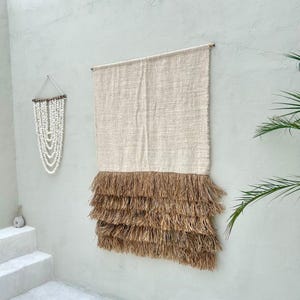 May include: A rectangular wall hanging featuring a woven beige fabric top section and a layered, fringed bottom section in a natural tan colour. To the left, a smaller wall hanging consists of multiple strands of white beads arranged in a V-shape.