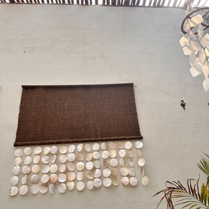 May include: A rectangular dark brown woven fabric wall hanging featuring multiple rows of circular, iridescent white shell discs suspended from the bottom edge, creating a layered, textured appearance against the plain wall surface.