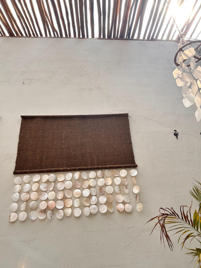 May include: A rectangular dark brown woven fabric wall hanging featuring multiple rows of circular, iridescent white shell discs suspended from the bottom edge, creating a layered, textured appearance against the plain wall surface.
