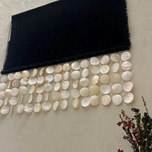 May include: A rectangular black fabric wall hanging featuring a horizontal row of dangling white iridescent circular shell discs. The textile has a fringed edge at the top and is suspended by a thin rod, with the shells arranged in multiple vertical strands.