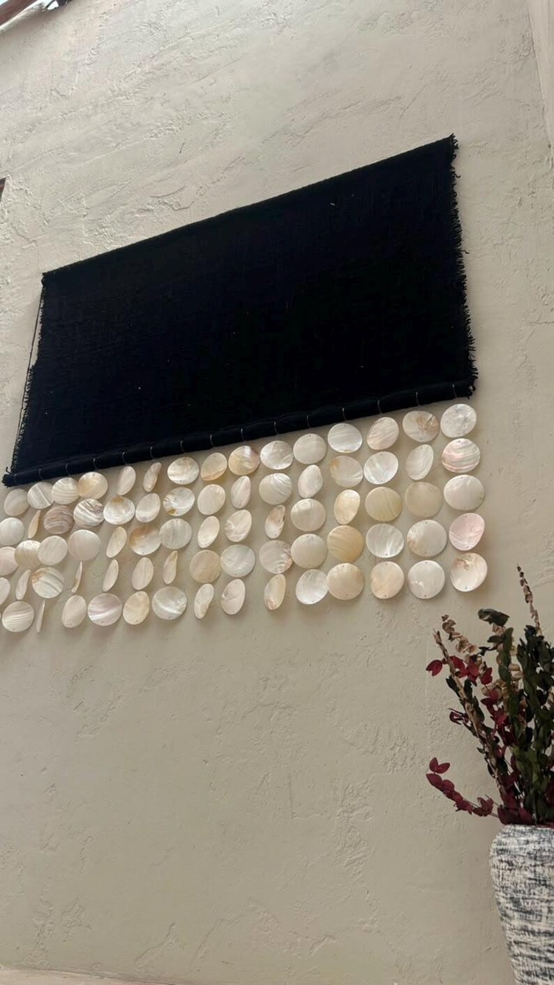 May include: A rectangular black fabric wall hanging featuring a horizontal row of dangling white iridescent circular shell discs. The textile has a fringed edge at the top and is suspended by a thin rod, with the shells arranged in multiple vertical strands.