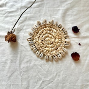 Puede incluir: Un posavasos redondo tejido de color beige natural con un borde de conchas blancas.