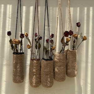 Könnte beinhalten: Fünf hängende Pflanzentöpfe aus gewebtem Material mit getrockneten Blumen. Die Halter sind zylindrisch, aus hellbraunem Material gefertigt und an Kordeln aufgehängt. Die Blumen sind eine Mischung aus Gelb und Rot.