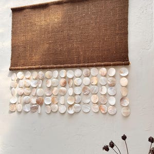 May include: A rectangular brown woven fabric wall hanging featuring multiple vertical strands of circular, iridescent white and cream-colored seashells suspended from the bottom edge of the textile in a grid-like arrangement.