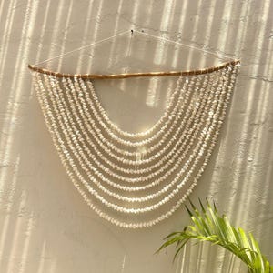 May include: A wall hanging featuring rows of white seashells suspended from a natural wooden branch. The shells are arranged in a curved, cascading pattern, creating a decorative and textured design. The piece is hung against a neutral-colored wall.