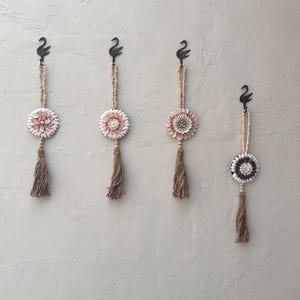 May include: Four decorative wall hangings. Each features a black swan hook, a beaded strand, a floral medallion with pink and white petals, and a brown tassel. The hangings are displayed against a neutral-colored wall.