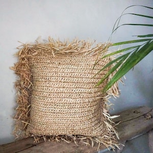 Puede incluir: Una almohada cuadrada tejida de color natural con un borde de flecos. La almohada está hecha de un material natural como la paja o la hierba marina.