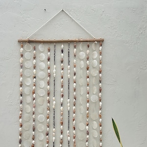 May include: A wall hanging featuring multiple vertical strands of small brown and white seashells interspersed with translucent circular capiz shell discs, all suspended from a horizontal wooden rod with a white cord for mounting.