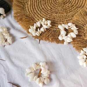 Puede incluir: Pendientes de conchas blancas sobre un fondo de tela blanca con dos posavasos de paja trenzada. Los posavasos tienen un borde de flecos.