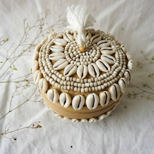 May include: A round, woven box with a white tassel on top. The box is decorated with white seashells and beads.