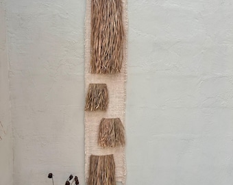 Può includere: Un arazzo da parete beige tessuto con quattro sezioni di materiale secco simile alla paglia. L'arazzo da parete è rettangolare e appeso verticalmente contro un muro grigio chiaro. Le sezioni di paglia sono di varie dimensioni e forme.