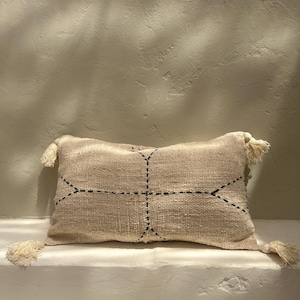May include: A beige throw pillow with black stitching in a geometric pattern. The pillow has tassels on each corner.