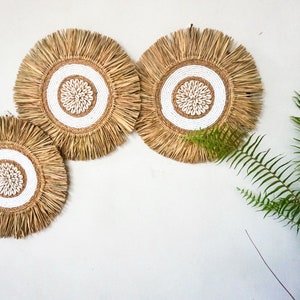 May include: Three round wall decor pieces made of natural materials. Each piece features a white circle with a smaller white circle in the center, surrounded by a ring of small white shells. The outer edge of each piece is made of a brown, fringed material.