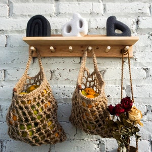Könnte beinhalten: Zwei braune Netztaschen gefüllt mit Orangen hängen an einem Holzregal mit Haken. Das Regal ist an einer weißen Backsteinwand montiert. Auf dem Regal befinden sich drei Dekorationsgegenstände, einer schwarz, einer weiß und einer braun.