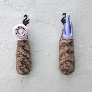 Puede incluir: Dos bolsas de almacenamiento colgantes de crochet marrón con ganchos de metal negro. La bolsa de la izquierda contiene un secador de pelo rosa. La bolsa de la derecha contiene un juego de herramientas de jardinería azules.