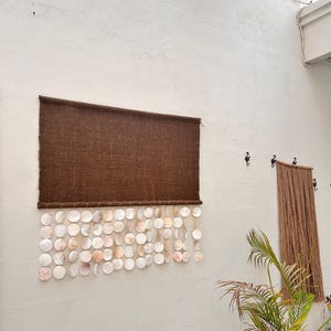 May include: A rectangular wall hanging featuring a dark brown woven fabric panel at the top, with multiple strands of circular, iridescent white and cream-colored shell discs suspended from the bottom edge in a layered, overlapping arrangement.