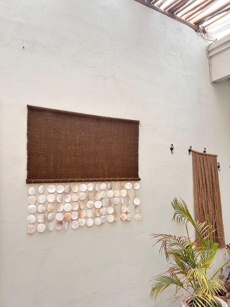 May include: A rectangular wall hanging featuring a dark brown woven fabric panel at the top, with multiple strands of circular, iridescent white and cream-colored shell discs suspended from the bottom edge in a layered, overlapping arrangement.