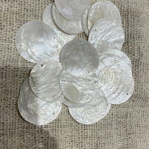 May include: A collection of circular, iridescent white shell discs, each featuring a small, singular hole near the edge. These flat, semi-translucent pieces display a natural, pearlescent sheen and are arranged in an overlapping cluster.